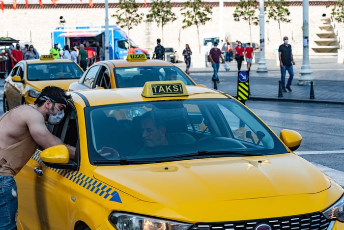 Gros plan sur un taxi jaune « TAKSİ » dans une rue animée, avec une personne penchée à la fenêtre côté conducteur et des piétons au fond.